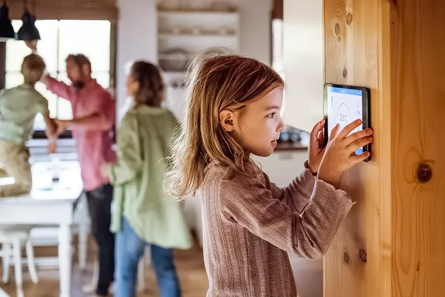 Welcher Stromverbrauch ist normal in einem privaten Haushalt? Ein Mädchen schaut sich den Stromverbrauch auf einem Tablet an, das an der Wand hängt, im Hintergrund sieht man verschwommen andere Familienmitglieder an einem Tisch reden